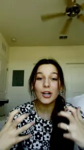 Young woman wearing a floral shirt, gesturing animatedly while speaking, with a bright indoor background and ceiling fan.