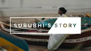 A woman in traditional attire sits beside colorful fishing boats, embodying resilience in Surubhi's Story.