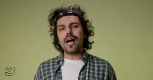 Man with curly hair and a bandana wearing a checkered shirt against a green background, smiling and speaking.