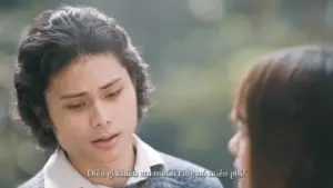 A young man with curly hair looks concerned while talking to a woman outdoors in a soft, natural setting.