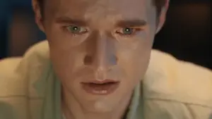 Close-up of a young man with green eyes, looking anxious and emotional while gazing at a computer screen in dim lighting.