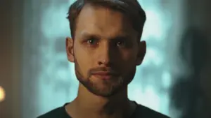 Close-up portrait of a man with a serious expression, soft lighting, and a blurred background creating atmospheric depth.