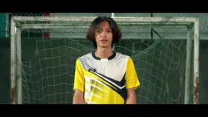 Young athlete in a yellow and white soccer jersey stands confidently in front of a goal net on a soccer field.