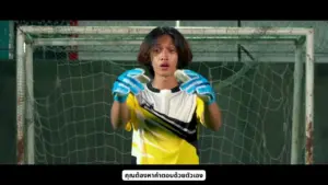 Young goalkeeper in a yellow jersey and blue gloves, preparing to defend a soccer goal in an indoor stadium.