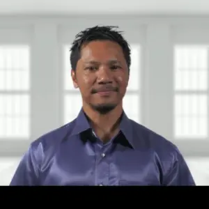 Smiling man in a blue shirt stands confidently in a bright, modern room with large windows.