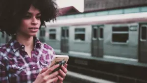 Young woman with curly hair using a smartphone at a subway station, train visible in the background.