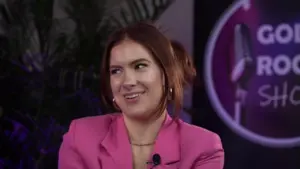 Woman in a pink blazer smiling at an event, with a microphone backdrop and vibrant lighting enhancing the atmosphere.