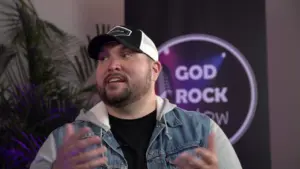 Man wearing a denim jacket and cap speaks animatedly in front of a "God Rock Show" backdrop with colorful lighting.