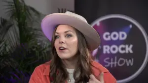 Woman in a wide-brimmed hat speaking passionately on stage with a colorful backdrop for a rock show event.