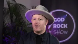 Man wearing a wide-brimmed hat, smiling against a colorful backdrop with "Good Rock Show" text and indoor plants.