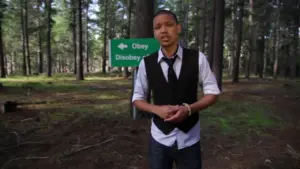 Man in a vest stands in a forest next to a sign pointing toward "Obey" and "Disobey" with tall trees in the background.