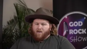 Man with curly hair and a hat speaking in front of a colorful backdrop, with a plant in the background.