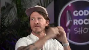 Man in a cap gestures while speaking on stage, surrounded by greenery and a modern backdrop at a music event.