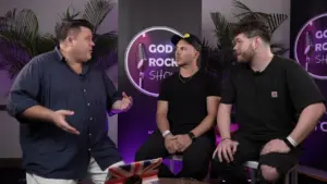 Three men engaged in conversation on a set with a vibrant backdrop, featuring a logo for the "God Rock Show."