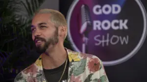 Man with blonde hair in a floral shirt speaking in front of a microphone backdrop for the God Rock Show.