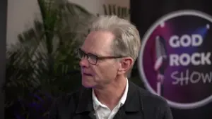 Man with gray hair, wearing a black jacket, engaged in conversation at a media event backdrop.