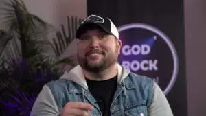 Smiling man in a denim jacket and cap, background with 'God Rock' graphic, vibrant atmosphere.