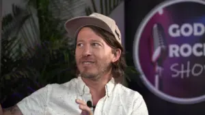 Man in a cap smiling and gesturing, with a microphone backdrop featuring the words "God Rock Show."