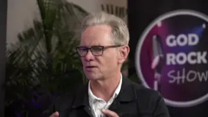 Man speaking passionately in front of a backdrop featuring the God Rocks Show logo, with greenery in the background.