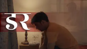 A young man in a yellow shirt sitting on a bed, reflecting in a dimly lit room with a bedside lamp.