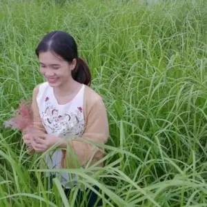 Young woman smiling in a lush green field, holding decorative grass, embodying joy and nature's beauty.