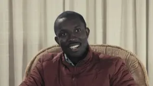 Smiling man in a red jacket seated in front of a beige curtain, exuding a friendly and approachable demeanor.