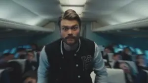 Man with a mustache in a varsity jacket standing in an airplane cabin, with passengers in the background.