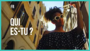 Young woman in sunglasses poses in a sunlit street, wearing a polka dot off-shoulder top, with urban buildings in the backdrop.