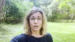 A woman with wavy hair and glasses stands outdoors, surrounded by lush greenery and trees in a bright, serene setting.