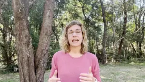 Woman with wavy hair standing outdoors, gesturing and speaking, surrounded by trees in a natural setting.