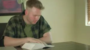 Man studying intently at a wooden table, with an open book and a relaxed indoor setting featuring soft lighting.