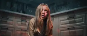 Young girl with long hair crouching on kitchen floor, looking scared in dimly lit room.