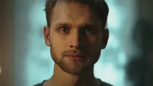 Close-up portrait of a man with a serious expression, illuminated by soft lighting, set against a hazy background.