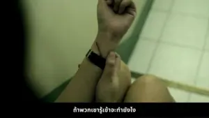 Person holding wrist with a watch, conveying emotion in a dimly lit space, with Thai text in the background.