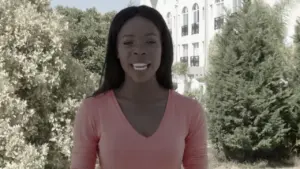 Smiling woman in a pink top stands outdoors, lush greenery and a building in the background, conveying warmth and professionalism.