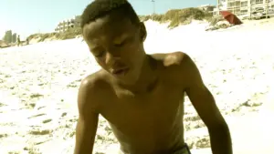 A young boy sitting on the sandy beach, focused on something in front of him under clear blue skies.