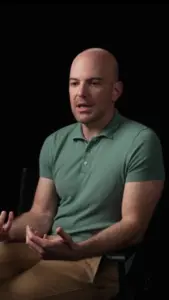 Man in a green polo shirt seated and speaking, with a black background enhancing the focus on his expressions.