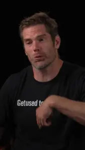 Man in a black t-shirt with text, gesturing while speaking, against a dark background. Focused expression.