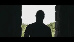 Silhouette of a person standing at a doorway, framed by stone walls, with lush greenery visible in the background.