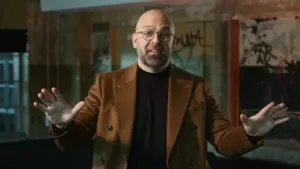 Man in a brown blazer gestures with hands, speaking confidently in an urban, graffiti-covered setting.