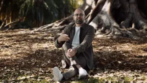 Man sitting on the ground among fallen leaves, gesturing with hand, in a serene outdoor setting under large trees.