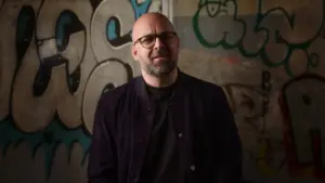 Man with glasses and a beard stands against a vibrant graffiti wall, exuding confidence and creativity.