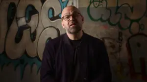 Man in glasses standing in front of a graffiti-covered wall, looking thoughtful and wearing a dark sweater.