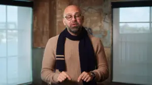 Man with glasses and a scarf stands in a well-lit room, thoughtfully gesturing while speaking to an audience.