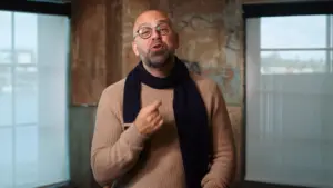 Person in a beige sweater and glasses gesturing while speaking in a rustic interior with windows and textured walls.