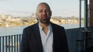 Professional man in a coastal setting, smiling, with a cityscape and water view in the background.