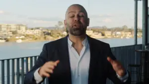 Man in a suit gestures passionately with a scenic waterfront view in the background, conveying energy and engagement.