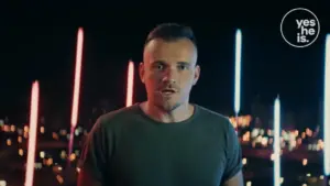 Man in a gray shirt with a surprised expression, illuminated by colorful neon lights in a nighttime cityscape.