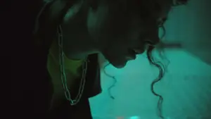 A person with curly hair leans down in low light, featuring a chain necklace and vibrant teal background.