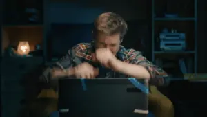 Person in a plaid shirt frustrated while working on a laptop in a dimly lit room, showcasing intense concentration.